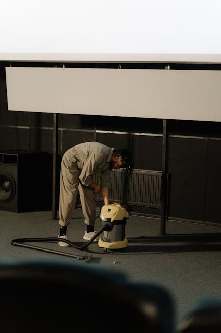 Close-up of a black vacuum cleaner head actively cleaning a patterned area rug with floral motifs in beige, green, and orange tones. The vacuum is positioned on the carpet, with a visible handle exten