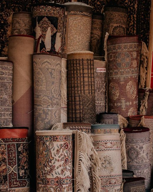 Close-up view of a decorative Persian-style rug with intricate geometric and floral patterns in red, orange, green, and beige hues, partially folded and resting on a stone or tiled floor. The rug appe