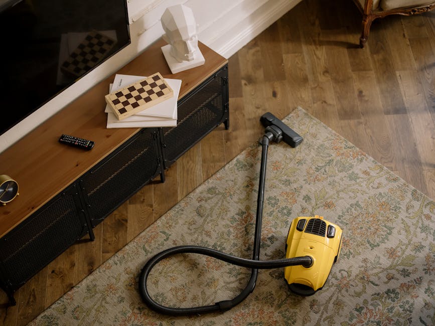 A yellow vacuum cleaner with a black hose and a metal wand attachment resting on an ornate patterned rug in a living room. In the background, there is a wooden TV stand with black metal legs, holding