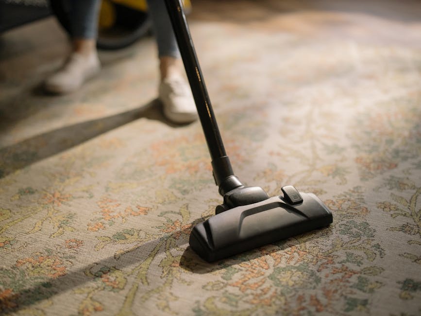 A yellow vacuum cleaner with a black hose and a metal wand attachment resting on an ornate patterned rug in a living room. In the background, there is a wooden TV stand with black metal legs, holding